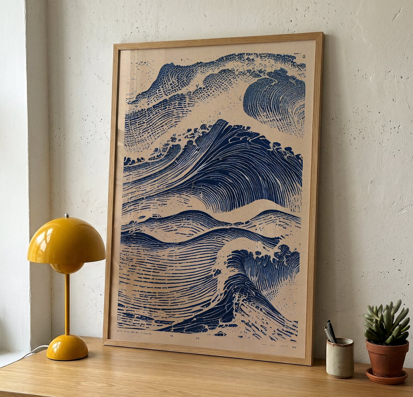 A framed art print affiche featuring a blue and beige wave illustration, leaning against a textured white wall on a wooden table next to a yellow lamp and a small potted succulent.