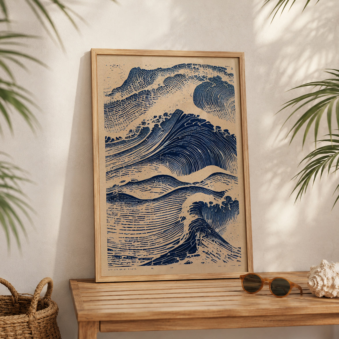 A framed art print featuring a blue and beige wave illustration, leaning against a wall on a wooden table with tropical leaves and sunglasses nearby.